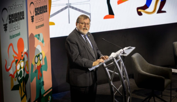 Prof Haymet stands at a lectern in front of a banner with National Science Week written on it