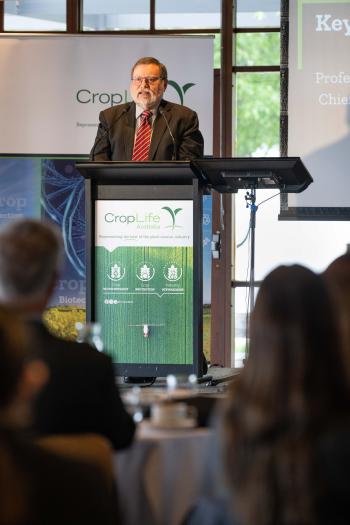 Prof Haymet stands at lectern on stage. Foreground shows attendees at tables