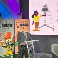 Prof Tony Haymet appears in front of a National Science Week banner, standing at a lectern. 